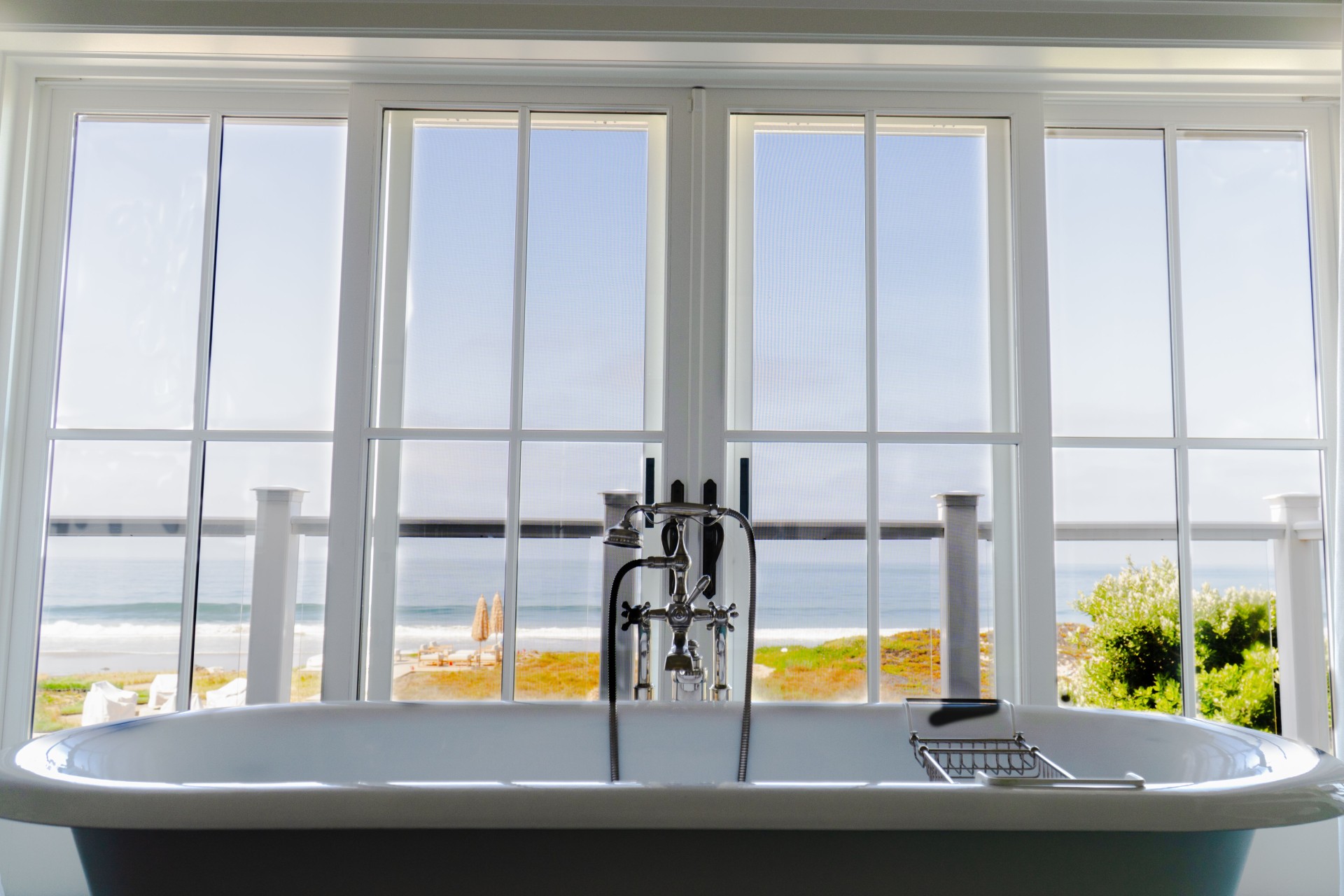 A white bathtub positioned in front of large windows overlooks a beach and ocean under a clear sky.
